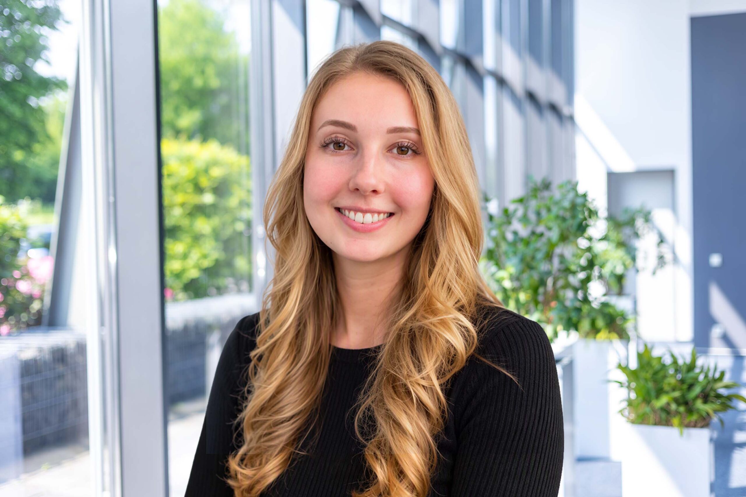 Lena Bock, Team Assistant, in front of large windows with green plants.