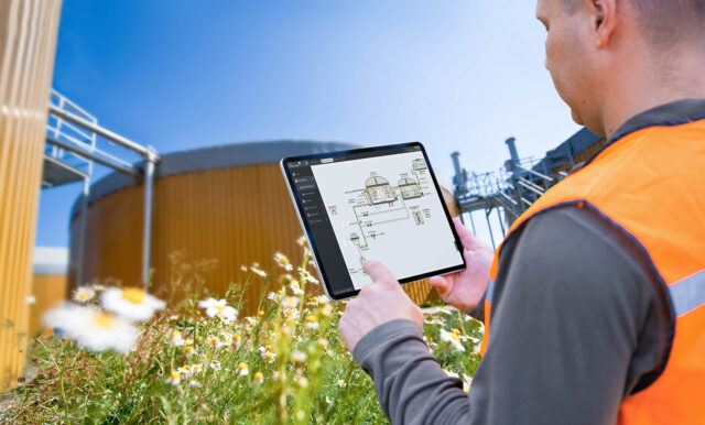 A person holds a tablet in front of an industrial facility outdoors.