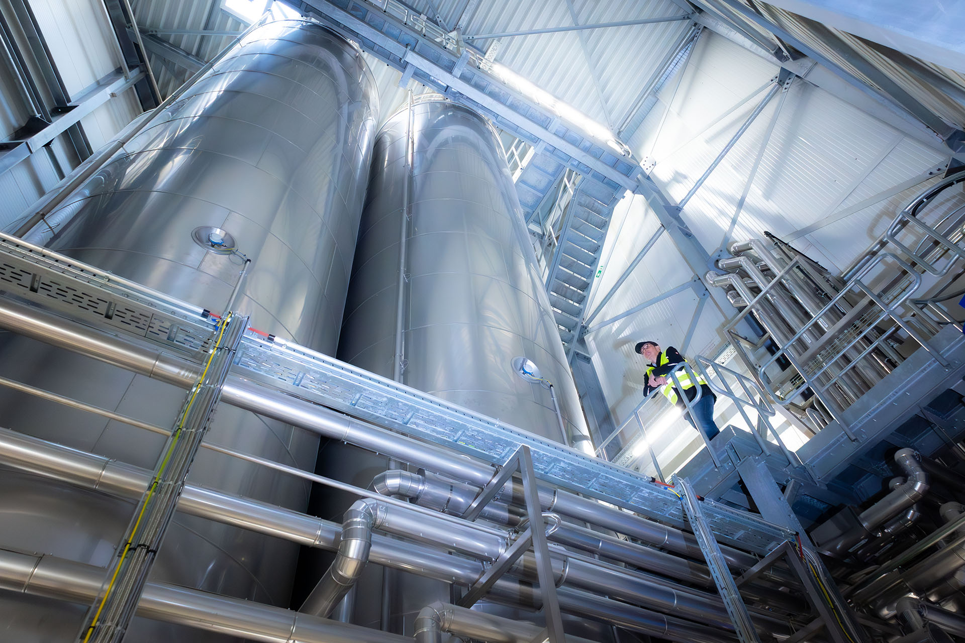 One person stands on a platform in front of large tanks and pipes.